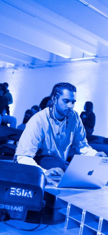 DJ sitting on the lounge floor, illuminated by blue light, mixing music on his laptop.