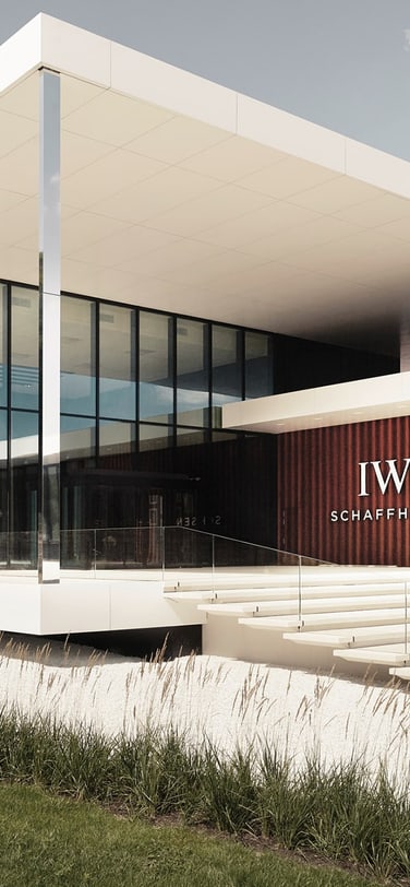 The Manufakturzentrum is a modern, two-story building with a flat white roof and large glass windows, featuring a textured wall with the "IWC Schaffhausen" logo.