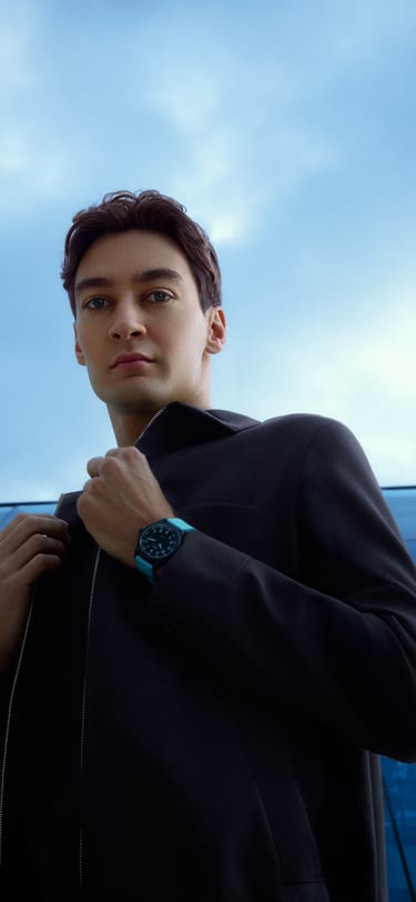 F1赛车手乔治·拉塞尔身着黑色赛车服，坐在玻璃建筑前，腕间佩戴一枚黑色陶瓷运动腕表，表带为蓝色。