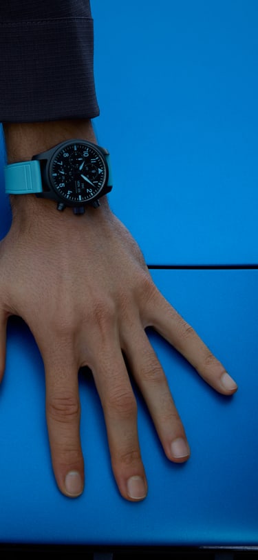 腕部特写，佩戴一块黑色陶瓷腕表，采用对比鲜明的蓝色表带、指针和时标，置于亮蓝色汽车引擎盖上，旁边即是银色AMG车标。