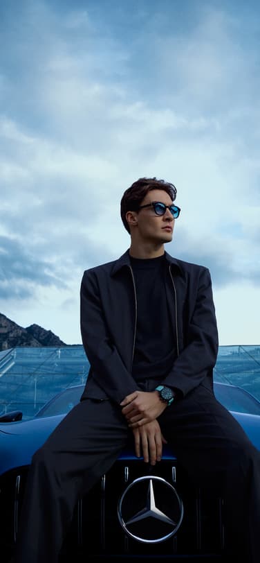 一级方程式赛车手乔治·拉塞尔身着深色服装，戴着蓝色眼镜，坐在梅赛德斯-奔驰车内，手腕上戴着一条蓝色表带的黑色陶瓷运动腕表，背景是摩纳哥城市、山脉和表镜建筑，天空多云。